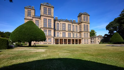 The image shows the lawn at Hardwick Hall, a prominent Elizabethan manor house located in Derbyshire, United Kingdom. Captured as an architectural landscape photograph, the scene is bathed in bright morning sunlight during the summer, with clear skies overhead. The historic Hardwick Hall, renowned for its symmetrical design and extensive windows, stands as a key National Trust property and well-known landmark in the region. The view highlights the manicured grounds and distinctive topiary, emphasizing the grandeur of Elizabethan architecture at this manor house in Derbyshire.